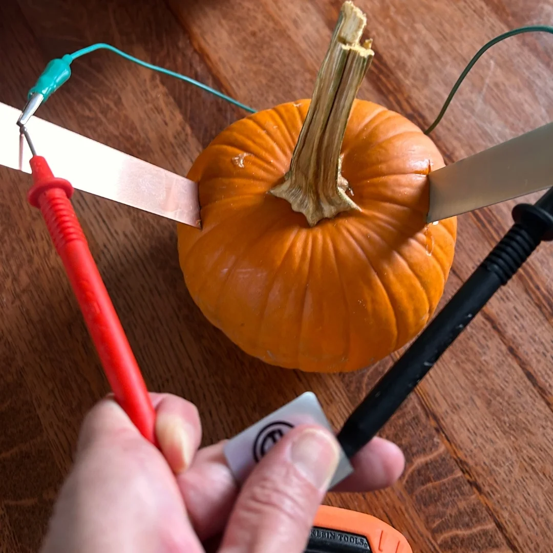 A small pumpkin with two metal strips inserted and wires attached demonstrates pumpkin power. A person holds red and black probes from a multimeter against the strips, testing how pumpkins can light a lightbulb on a wooden surface.