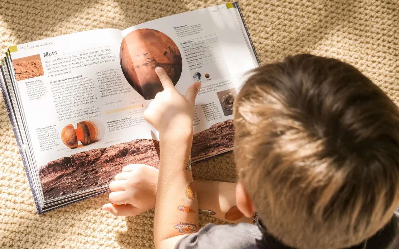 A child is lying on a textured carpet, absorbed in reading an open book about Mars. They point at the planet's image, colorful space-themed stickers adorning their arm. Sunlight illuminates this summer science exploration.