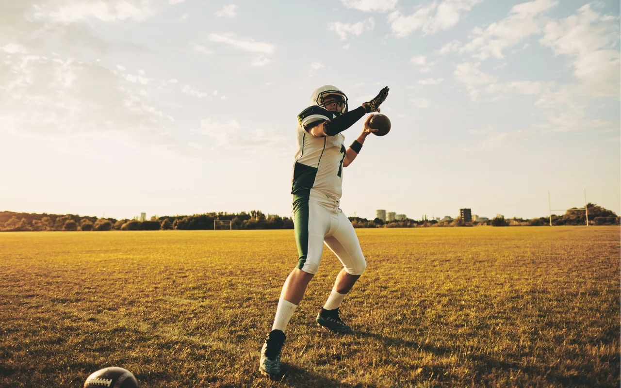 A football player wearing a green and white uniform with a helmet stands on a grassy field ready to throw a football. The sun is setting, casting a golden light over the scene with a city skyline visible in the distance. Another football lies on the ground nearby.
