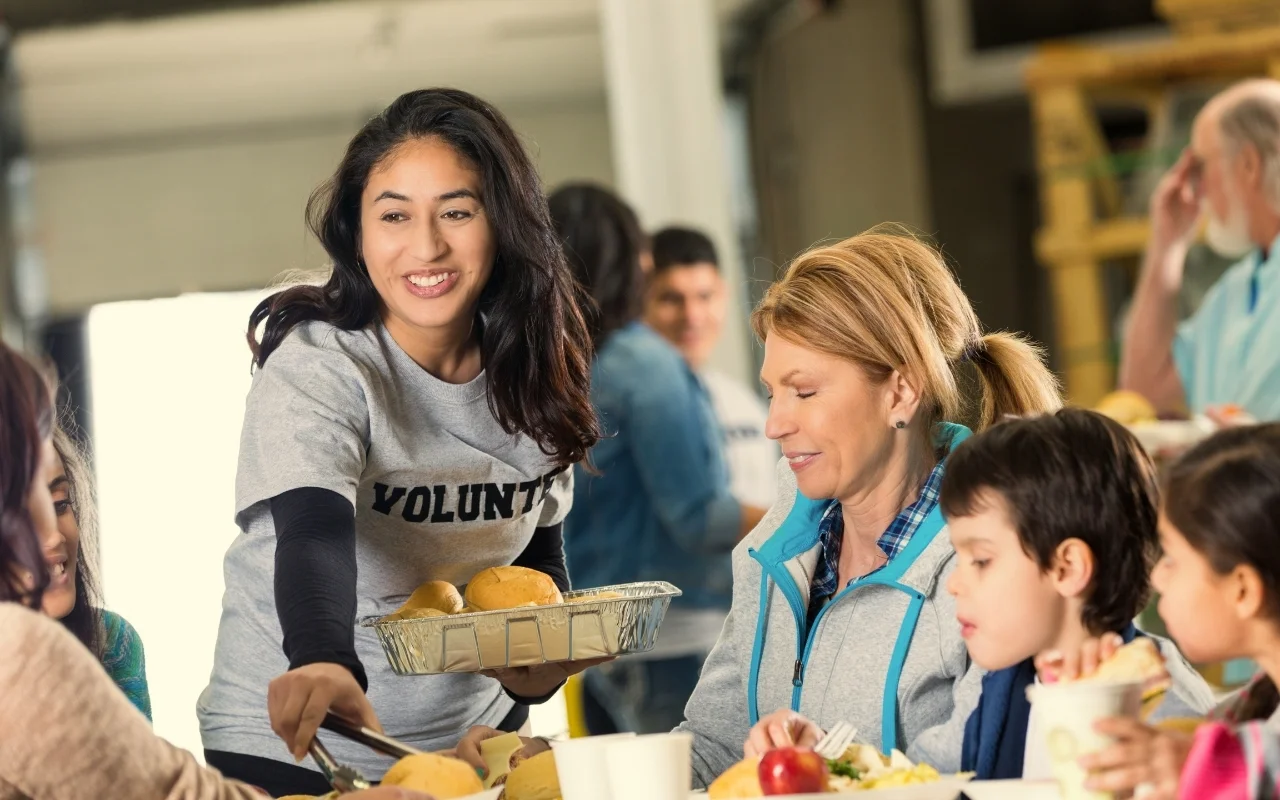 A smiling woman in a "Volunteer" shirt serves bread rolls at a community meal, inspiring a warm atmosphere where giving back encourages a positive mindset change for everyone involved.