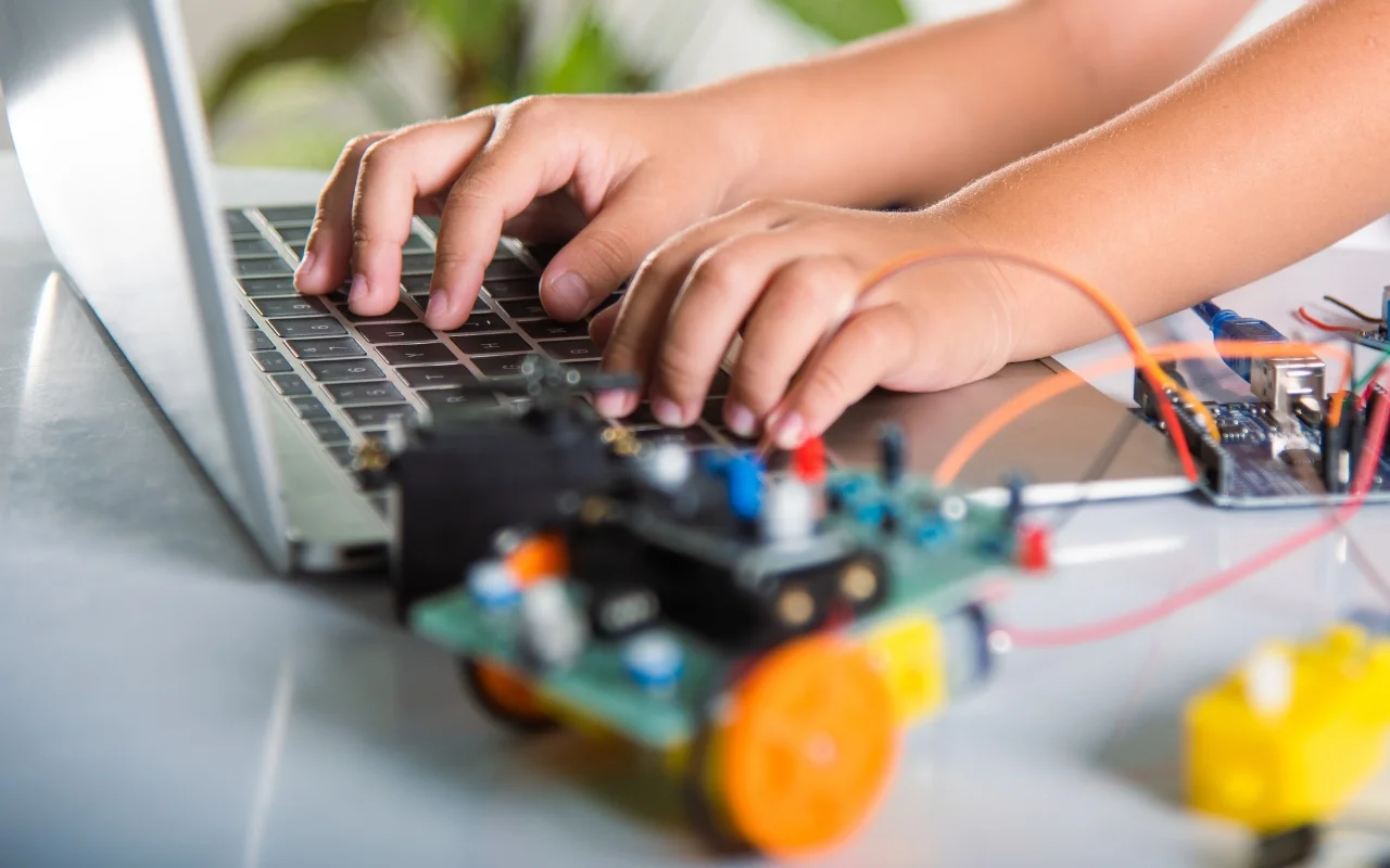 Hands typing on a laptop with a partially visible robotics project in the foreground, featuring a circuit board and wires. The scene captures a moment of discovery and curiosity as ideas come to life through organized innovation.