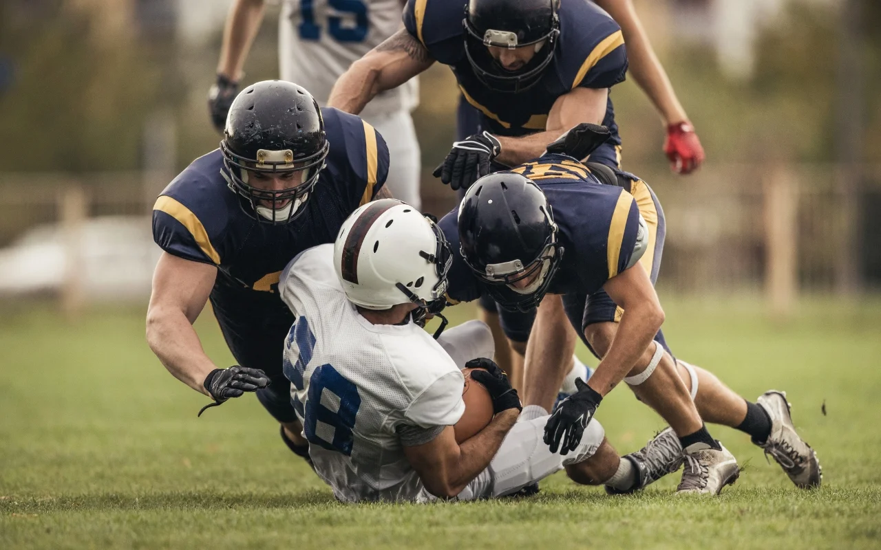 Three football players in black and yellow uniforms tackle a player in a white and blue uniform on a grassy field. The tackled player is holding the ball tightly as the others converge on him. The action is intense, with visible motion and determination.