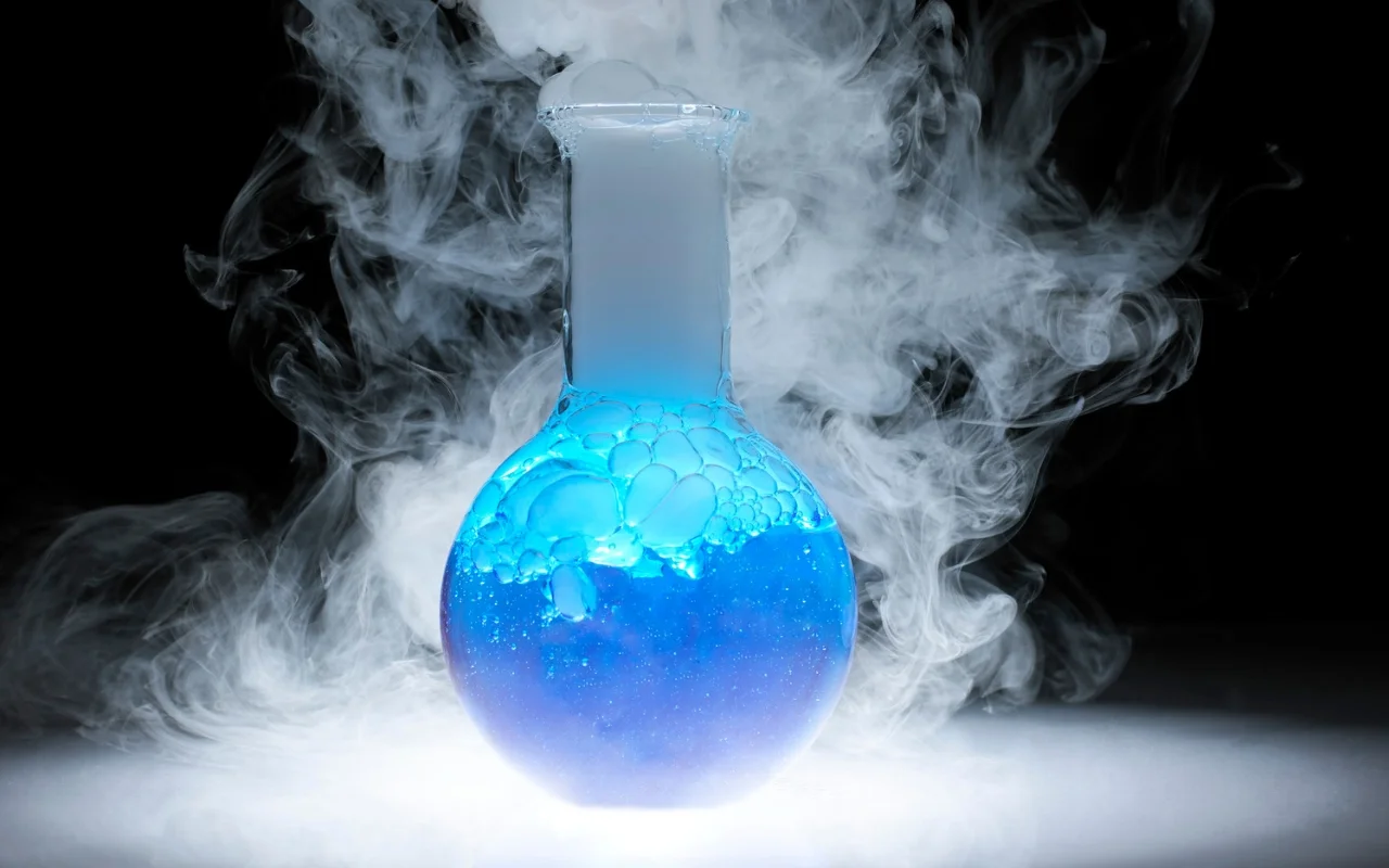 A round-bottom flask filled with bubbling blue liquid and dry ice emits thick white smoke, set against a dark background. The swirling smoke creates a mysterious and scientific atmosphere, perfect for shaking off the winter blahs with exciting science experiments.