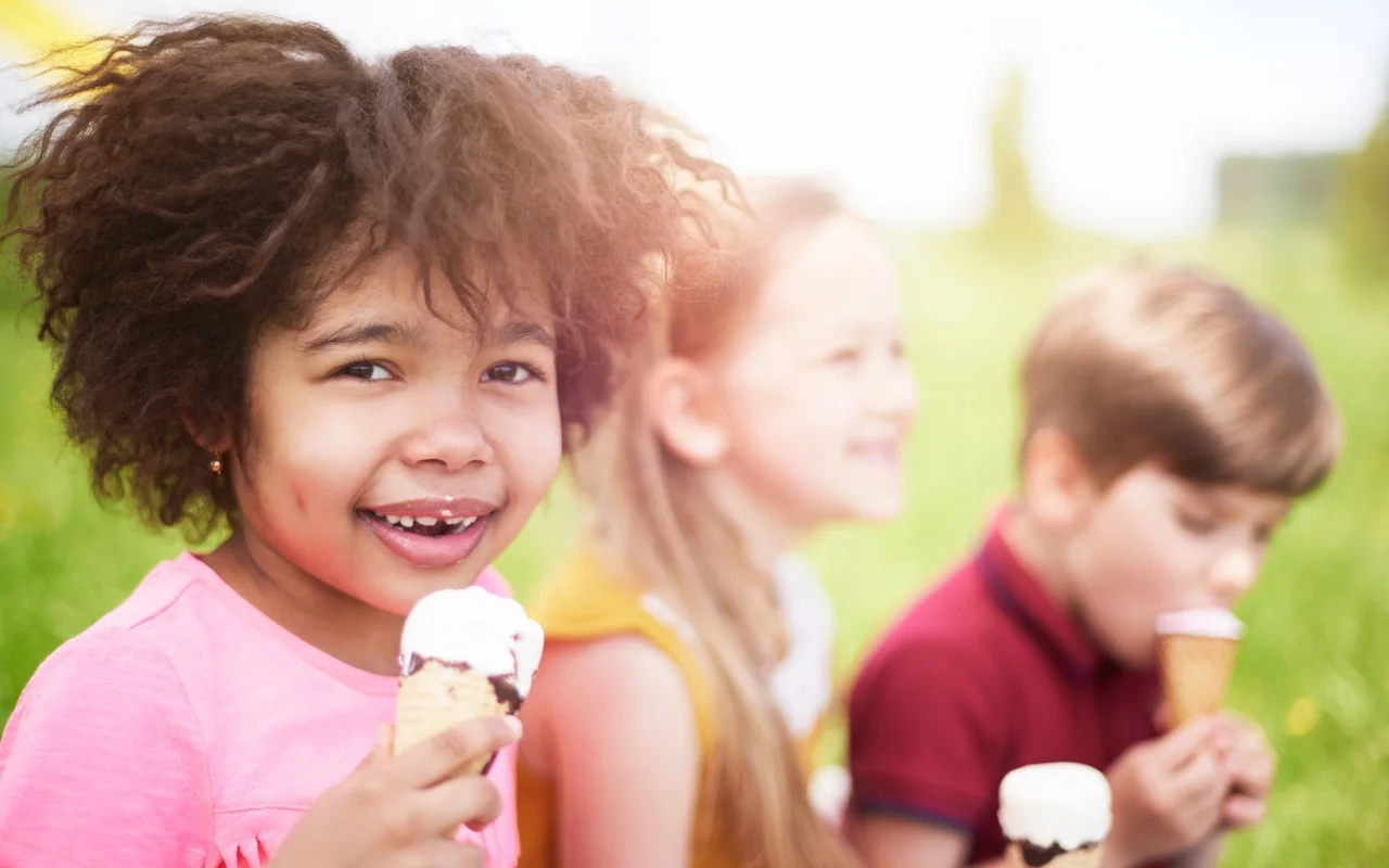 Three children enjoying ice cream cones outdoors. A girl in a pink shirt smiles at the camera, capturing the essence of summer fun, while the other two are blurred in the background, savoring their ice cream. The setting is a sunny, grassy area perfect for exploration.