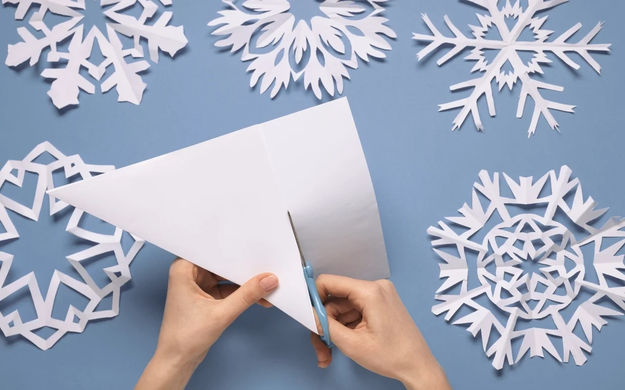 Hands cutting a folded piece of white paper with scissors to make a six-sided snowflake, surrounded by completed paper designs that showcase beautiful snowflake symmetry on a light blue background.