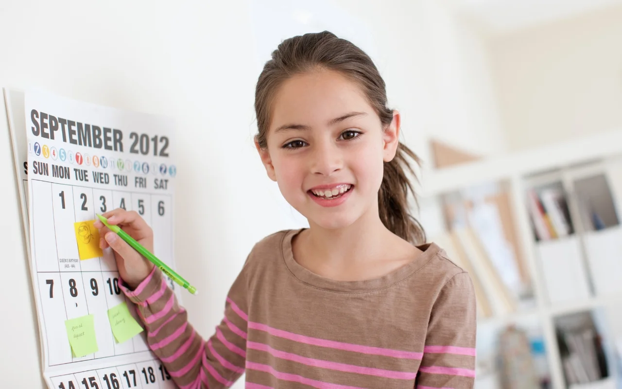 A smiling girl in a brown striped shirt marks a date on a wall calendar for September 2012, using her homeschool year planning. The calendar, dotted with sticky notes, hangs in a bright room with shelves in the background.