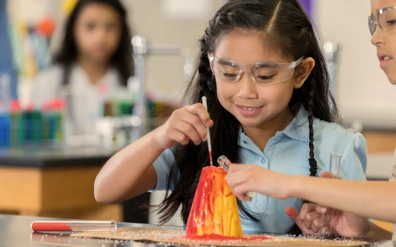 Two young girls wearing safety goggles conduct a science experiment in a classroom. They use a pipette and a test tube to create a volcanic eruption model with red and yellow lava on the table. In the background, lab equipment and another student are visible.