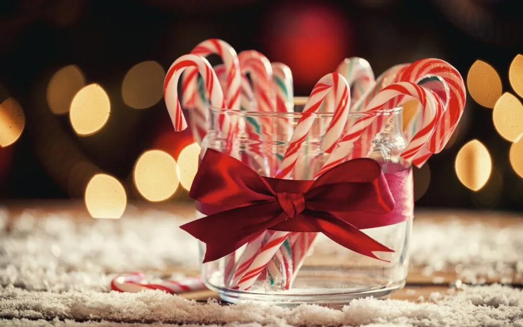 A glass jar filled with red and white candy canes, decorated with a red bow, sits on a snow-like surface. Warm, soft lights create a festive holiday atmosphere in the blurred background.